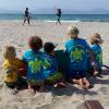 Cinq enfants, avec des t-shirts Horizon Surfing, qui jouent avec une planche de surf sur la plage face à la mer avec deux kitesurfeurs devant 