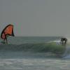 Spot de surf, kitesurf et wingfoil à Lassarga ION Club avec un surfeur sur une vague creuse et un wing derrière sur une eau bleu claire turquoise
