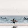 Spot de surf à Dakhla Lassarga avec des bâteaux de pécheurs au loin, un surfeur et sa longboard, des mouettes dans le ciel et du sable du désert du Sahara